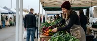 Young woman vendor arranging fresh organic vegetables at farmers market stall. Local business owner selling healthy produce outdoors.
