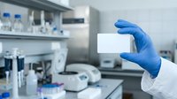Scientist wearing blue latex gloves holding blank white card in modern laboratory with equipment and samples on workbench for research, medical, or pharmaceutical applications.