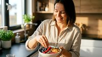 Happy Asian woman enjoying healthy breakfast bowl with fresh berries and granola in modern kitchen during morning sunlight.