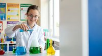 Young Caucasian female scientist in white lab coat and safety glasses conducting chemistry experiments with colorful solutions in laboratory setting.