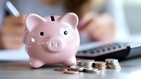 Pink ceramic piggy bank with stack of coins and calculator in background, representing financial savings, budgeting and money management concept.