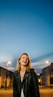 Young woman with shoulder-length auburn hair, Caucasian ethnicity, wearing black leather jacket over white shirt, smiling outdoors at dusk with street lights and buildings.