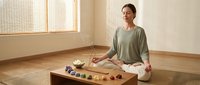 Young Asian woman in green top practicing meditation in minimalist room with wooden floor, natural light, and healing stones on table for wellness.