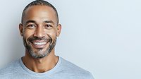Confident mature African American man with salt and pepper beard wearing grey t-shirt, displaying genuine warm smile against light background. Professional headshot style portrait.