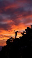 Silhouette of person with raised arms celebrating success on mountain peak against dramatic orange and purple sunset sky with clouds for motivation concepts.