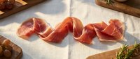 Thinly sliced prosciutto ham arranged on white linen with wooden cutting boards and herbs in warm natural sunlight for gourmet cooking and dining.