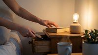 Woman hands opening wooden bedside table drawer in cozy bedroom with warm lamp light creating peaceful nighttime atmosphere for relaxation.