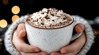 Cozy hands holding white mug with hot chocolate topped with whipped cream and chocolate sprinkles against blurred warm bokeh lights background.