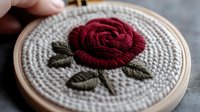 Handcrafted embroidery featuring deep red rose with olive green leaves on white fabric in wooden hoop, showcasing detailed needlework and traditional craft technique.
