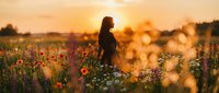 Woman standing in blooming wildflower field at golden hour sunset, surrounded by red poppies and white flowers, warm evening light creating bokeh background effect.