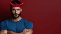 Bearded muscular man in blue t-shirt wearing red devil horns headband with crossed arms against dark red background, looking serious and confident.