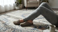 Woman relaxing at home in gray leggings and suede slippers on a vintage patterned rug, cozy living room lifestyle scene.
