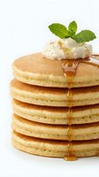 Stack of golden fluffy pancakes with maple syrup dripping down sides, topped with whipped cream and fresh mint leaves on white background.