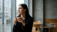 Young professional woman enjoying morning coffee while gazing thoughtfully out window in modern cafe setting with natural lighting and urban view.