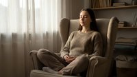 Woman with long dark hair and light skin tone, wearing glasses and a beige knit sweater, sitting cross-legged in a comfortable armchair by a window with natural light streaming through sheer curtains, relaxed home environment with bookshelves in background.