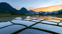 Terraced rice paddies reflect golden sunset sky and mountain silhouettes, creating geometric patterns in flooded fields during cultivation season in Asia.
