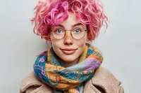 Young woman with pink curly hair, round glasses and colorful knit scarf smiling at camera against light background. Modern fashion portrait expressing joy and style.