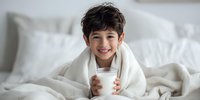 Happy young boy with dark hair wrapped in white blanket holding glass of milk in bright bedroom, smiling at camera with cozy morning atmosphere.