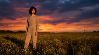 Woman with curly dark hair wearing beige blazer standing in golden wheat field at sunset. Outdoor fashion portrait for editorial, lifestyle, and commercial design use.