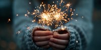 Hands holding bright sparklers creating magical golden sparks and light trails against soft blue background for celebration and festive occasions.
