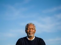 Senior man with gray hair and dark skin, wearing a dark ribbed sweater, smiling at camera against blue sky background. Suitable for healthcare, wellness, lifestyle and diversity concepts.