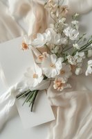 Delicate white and peach cosmos flowers with blank card on soft silk fabric background, creating romantic wedding stationery flatlay composition.