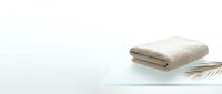 Folded beige towel on glass shelf with palm leaf and white background, spa and bathroom decor for wellness design projects.