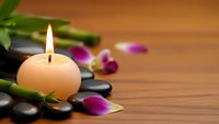 Spa wellness scene with burning candle, bamboo stems, purple orchid petals and black stones on wooden background for relaxation and meditation.