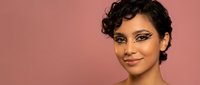 Young mixed-race woman with curly hair and dramatic eye makeup smiling confidently against pink background for beauty and fashion campaigns.