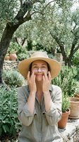 Woman in straw hat applying sunscreen in olive grove garden with terracotta pots, skincare and sun protection concept for outdoor beauty and wellness campaigns.