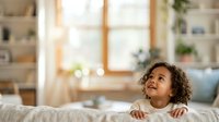 Young child with curly hair and medium skin tone standing at furniture edge, looking upward with curiosity in bright, sunlit living room with shelving and windows.
