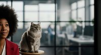 Woman with afro hairstyle wearing red blazer posing with gray tabby cat in modern office with city skyline view through windows.