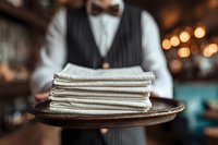 Elegant waiter in formal attire holding wooden tray with stack of folded white napkins in upscale restaurant with warm ambient lighting.