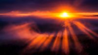 Dramatic sunset above clouds with golden rays breaking through dark storm clouds, creating atmospheric light beams for inspirational and nature backgrounds.
