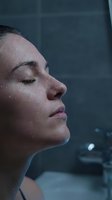 Woman with wet skin and closed eyes in profile, standing under shower with water droplets on face, relaxing in bathroom with tiled walls, wellness and self-care concept.