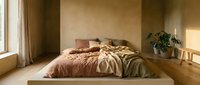 Minimalist bedroom with platform bed, terracotta and olive bedding, potted plant, wooden stool, and warm natural light from window for interior design projects.