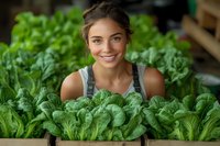 Young woman with brown hair and fair complexion, wearing denim overalls, smiling while surrounded by fresh green lettuce in a vegetable garden or farm setting.