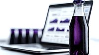 Purple beverage bottle with laptop displaying business charts in background, selective focus on glass container against blurred modern workspace setup.