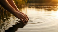 Hand touching calm water surface creating ripples at golden hour sunset, suitable for wellness, mindfulness, and nature-themed projects.
