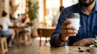 Man in blue shirt holding white coffee cup at wooden table in cafe interior with blurred background, suitable for beverage, lifestyle and business concepts.