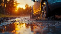 Car driving through muddy puddle at sunset creating dramatic splash and reflection in water. Dynamic low angle view captures motion and golden hour atmosphere.