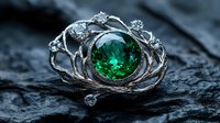Ornate silver ring with large emerald green gemstone nestled in intricate floral metalwork design, photographed on dark textured stone surface in dramatic lighting.