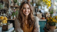 Young Caucasian woman with long wavy brown hair wearing mustard sweater and cardigan smiling at camera in cozy flower shop interior with yellow blooms and potted plants.