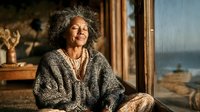 Smiling mature African American woman with curly gray hair in cozy sweater relaxing at home by window with warm natural lighting for lifestyle content.