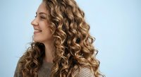 Young woman with voluminous curly blonde hair smiling in profile against light blue background. Natural beauty portrait for haircare and styling concepts.