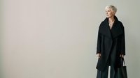 Mature woman with short gray hair, Caucasian ethnicity, wearing black formal coat and dress, standing against neutral background with copy space for business or corporate use.