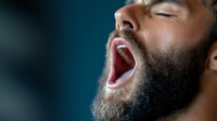 African American man with beard yawning or expressing tiredness against dark background, close-up portrait showing natural emotion and facial expression.