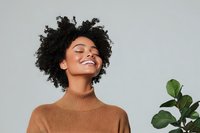 Young African American woman with natural curly hair wearing brown turtleneck sweater expressing joy and contentment against gray background with green plant leaf.