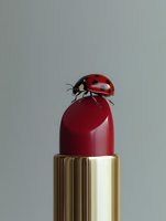 Red ladybug perched on burgundy lipstick tube with golden case against neutral background for beauty and cosmetics advertising campaigns.