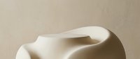 Abstract beige ceramic sculpture with smooth organic curves against a neutral textured wall, evoking minimalist art and modern interior decor.
